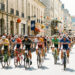 Peloton de cyclistes hommes et femmes traversant une ville française en été, avec une foule dense derrière les barrières et des décorations colorées.