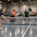 Deux femmes et deux hommes jouent au pickleball sur un terrain intérieur lumineux près d’Arras, avec une balle perforée visible.