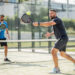 Deux hommes amateurs de padel en double se préparent pour un service sur un terrain vitré, dans une ambiance lumineuse et réaliste.